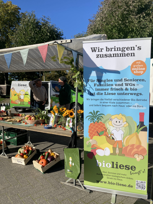 KI generiert: Ein Marktstand mit Obst, Gemüse und einem Werbebanner der Bioliese für Bio-Produkte und Onlineshop.