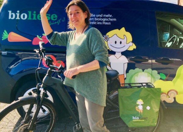 KI generiert: Frau mit Fahrrad vor einem Lieferwagen mit "bioliese"-Logo. Text: "bioliese bringt's!"
