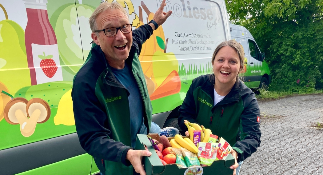 KI generiert: Zwei Personen halten eine Kiste mit Obst und Gemüse vor einem Lieferwagen. Text: "biofrische".