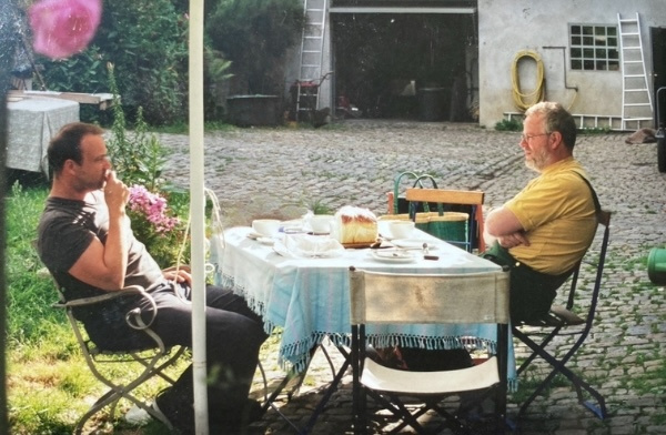KI generiert: Zwei Männer sitzen im Garten an einem Tisch mit gedecktem Frühstück. Hintergrund: gepflasterter Hof und Garage.