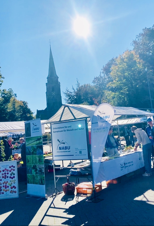 KI generiert: Marktstand von NABU mit Infomaterial, umgeben von Menschen. Text: "Unterstützen Sie uns! Gemeinsam für Mensch und Natur."