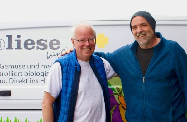 KI generiert: Zwei Männer stehen vor einem Lieferwagen mit Werbung für "Obst, Gemüse und mehr aus kontrolliert biologischem Anbau."