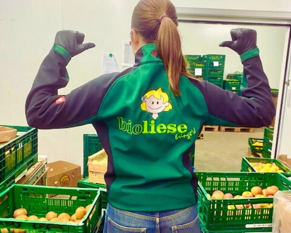 KI generiert: Eine Person zeigt auf ihre "bioliese bringt's!"-Jacke, umgeben von Kisten mit Gemüse.