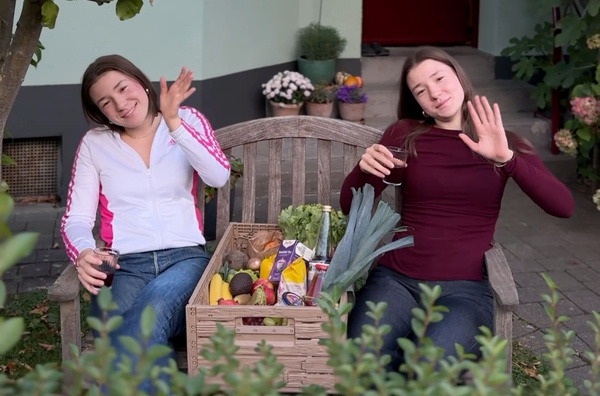 KI generiert: Zwei Personen sitzen mit Getränken und einer Kiste voller Obst und Gemüse auf einer Bank und winken.