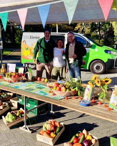 KI generiert: Ein Marktstand mit Obst und Gemüse, drei Personen, und ein Lieferwagen mit "bioliese" Schriftzug.