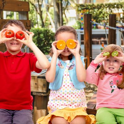 KI generiert: Drei Kinder halten Obst und Gemüse vor ihre Augen und sitzen auf einer Bank.