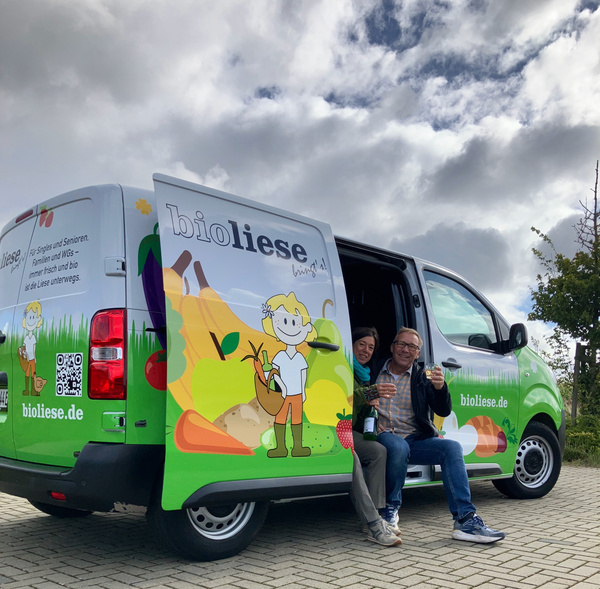 KI generiert: Ein bioliese-Lieferwagen mit bunter Grafik und zwei lachende Personen sitzen in der geöffneten Tür. Text: "bioliese.de".