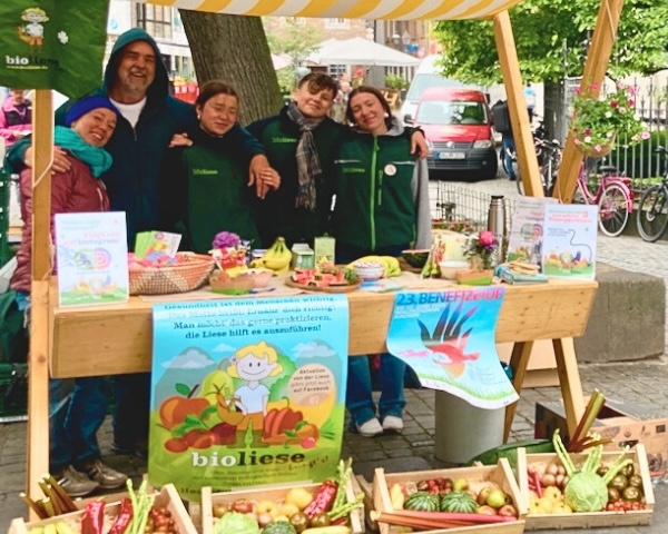 KI generiert: Eine Gruppe von Menschen an einem Marktstand mit Gemüse und Postern. Texte: "bioliese", "23. BENEFIZ".
