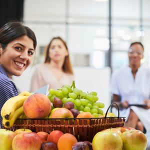 KI generiert: Frau lächelt neben einem Korb voller Obst, darunter Bananen, Trauben und Äpfel.
