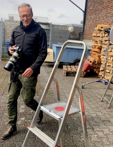 KI generiert: Ein Mann mit Kamera neben einer Aluminiumleiter im Freien, umgeben von gestapeltem Holz.