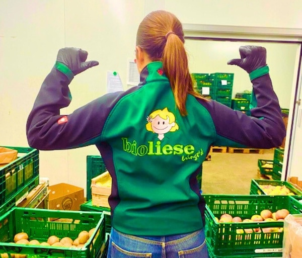 KI generiert: Frau zeigt auf Rücken, trägt Jacke mit Text: "bioliese bringt’s!"; umgeben von grünen Kisten mit Obst/Gemüse.