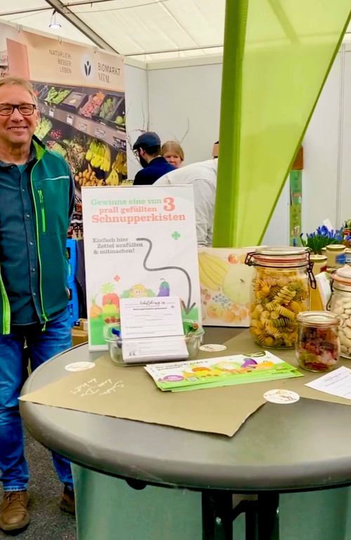 KI generiert: Ein Messestand mit Bio-Produkten und einem Gewinnspiel. Text: "Gewinne eine von 3 prall gefüllten Schnupperkisten."