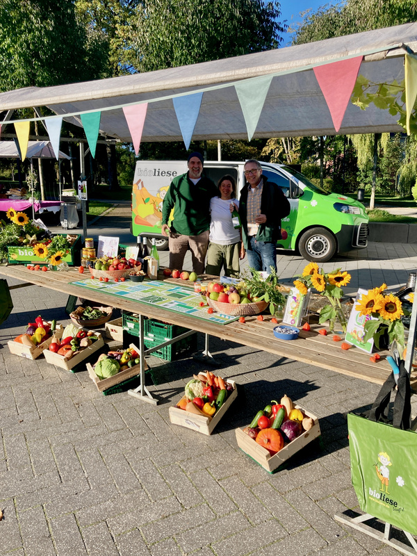 KI generiert: Marktstand mit Gemüse und Obst, drei Personen, farbige Wimpel, Sonnenblumen, Aufschrift: "bioliese".
