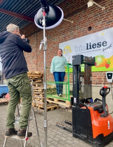 KI generiert: Ein Fotograf macht ein Foto von einer Person vor einem "bioliese"-Banner mit einem Gabelstapler im Vordergrund.