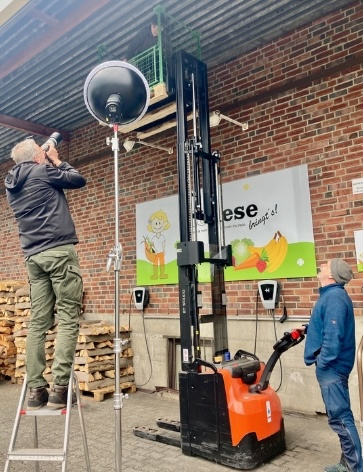 KI generiert: Zwei Personen bei einem Fotoshooting in einem Lager, einer auf einer Leiter, ein anderer mit Gabelstapler.